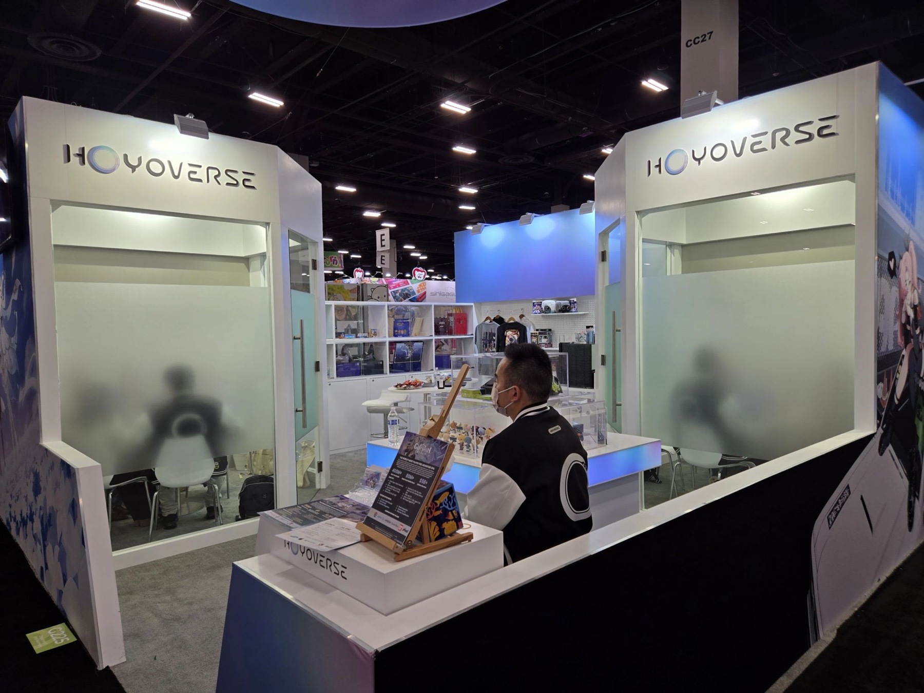 Wide shot of the HoYoverse booth at Licensing Expo 2025, with frosted glass meeting rooms, the brand logo across both sides, and a rep at the front desk.