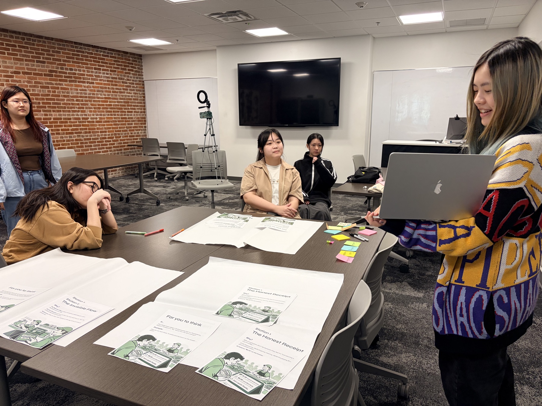 Workshop participants seated around a table with station-card printouts and sticky notes